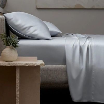 Bed with light gray sheets and pillows, a small plant in a vase on a stone side table.