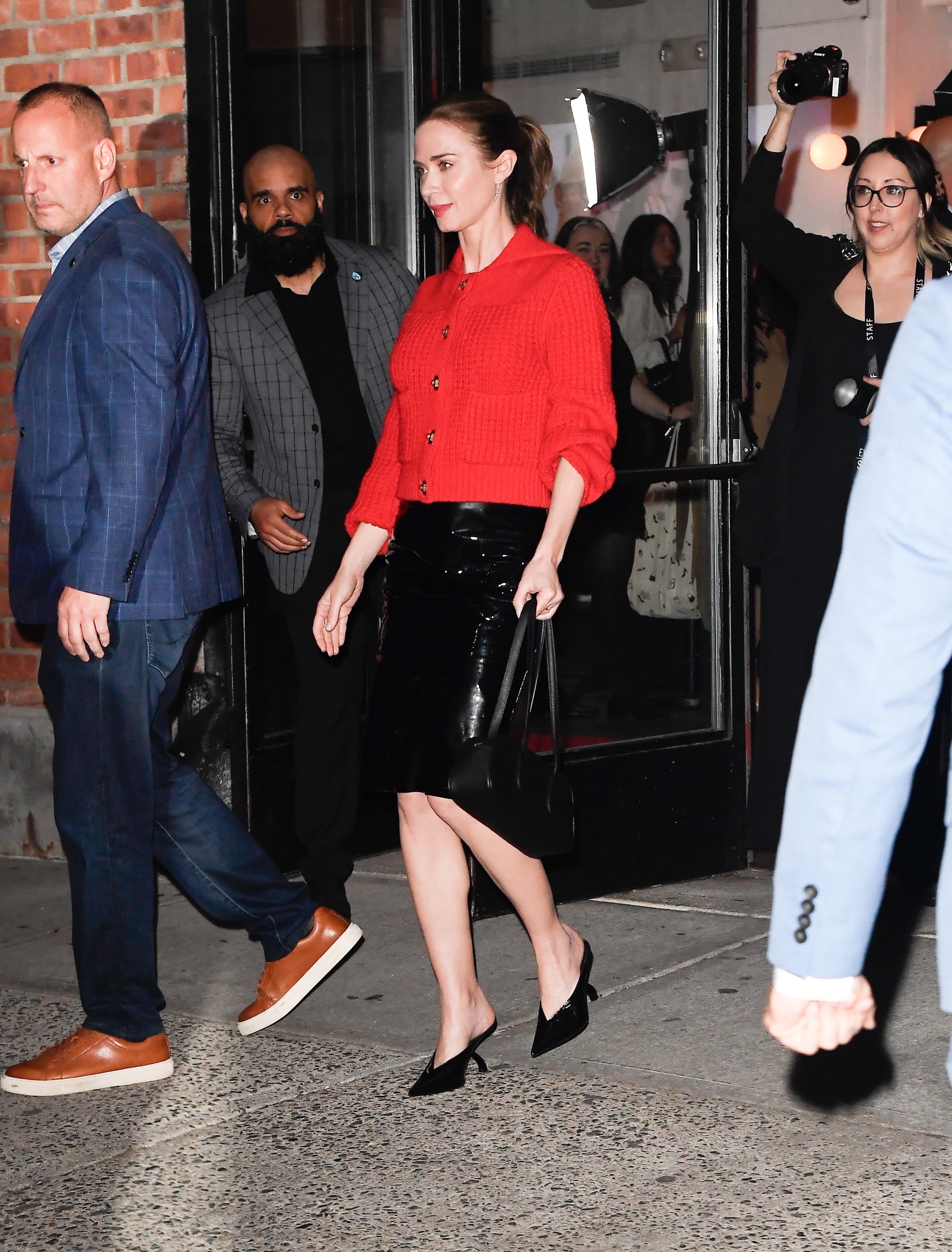 Emily Blunt in a red sweater and black leather skirt holding a baguette bag while promoting "The Devil Wears Prada 2" in New York City.